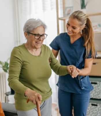 A caregiver assists an elderly woman with walking inside a home.